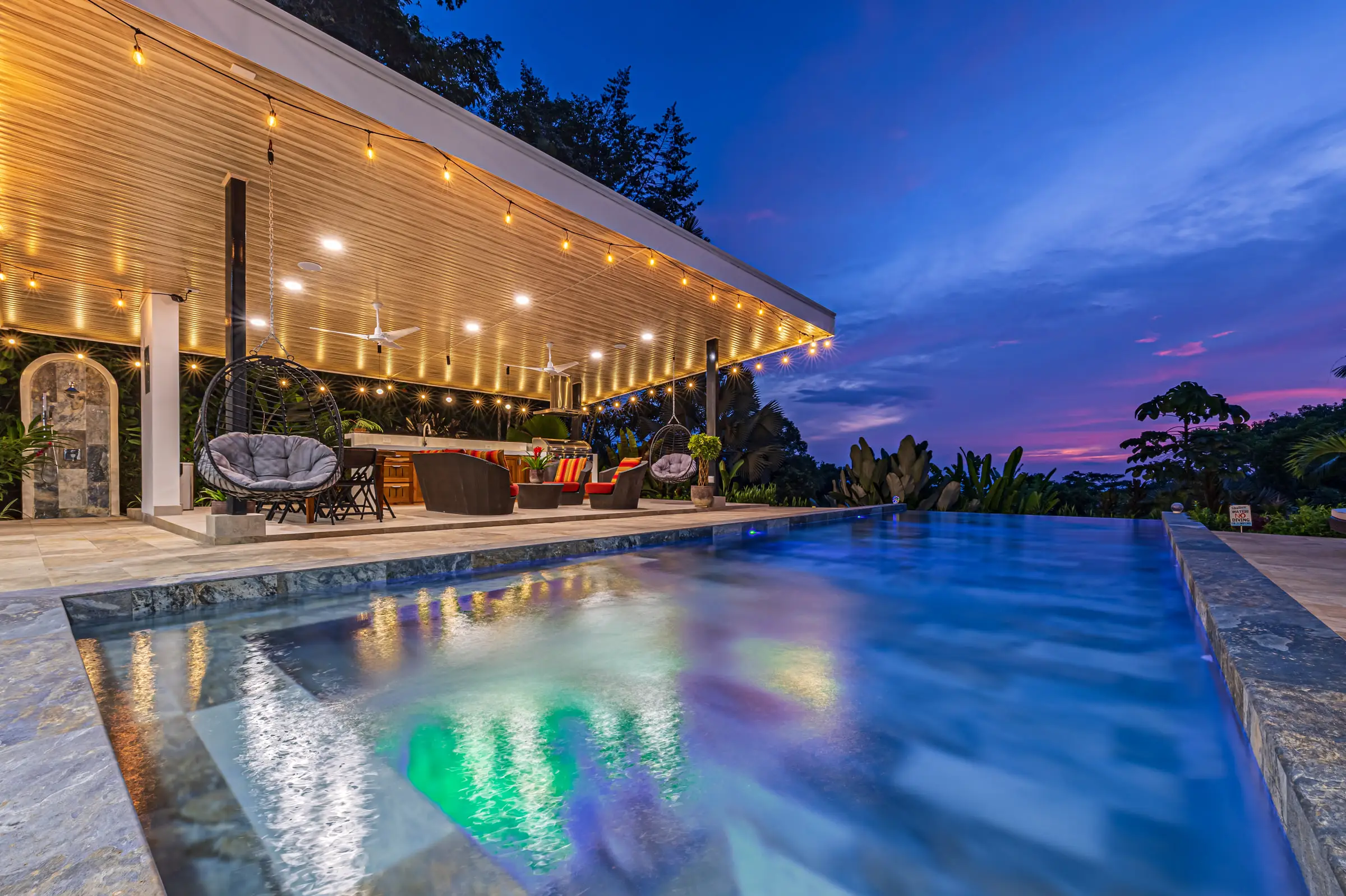 The infinity pool at dusk, lit from below, with string lights along the covered outdoor kitchen and a violet sky over the forest